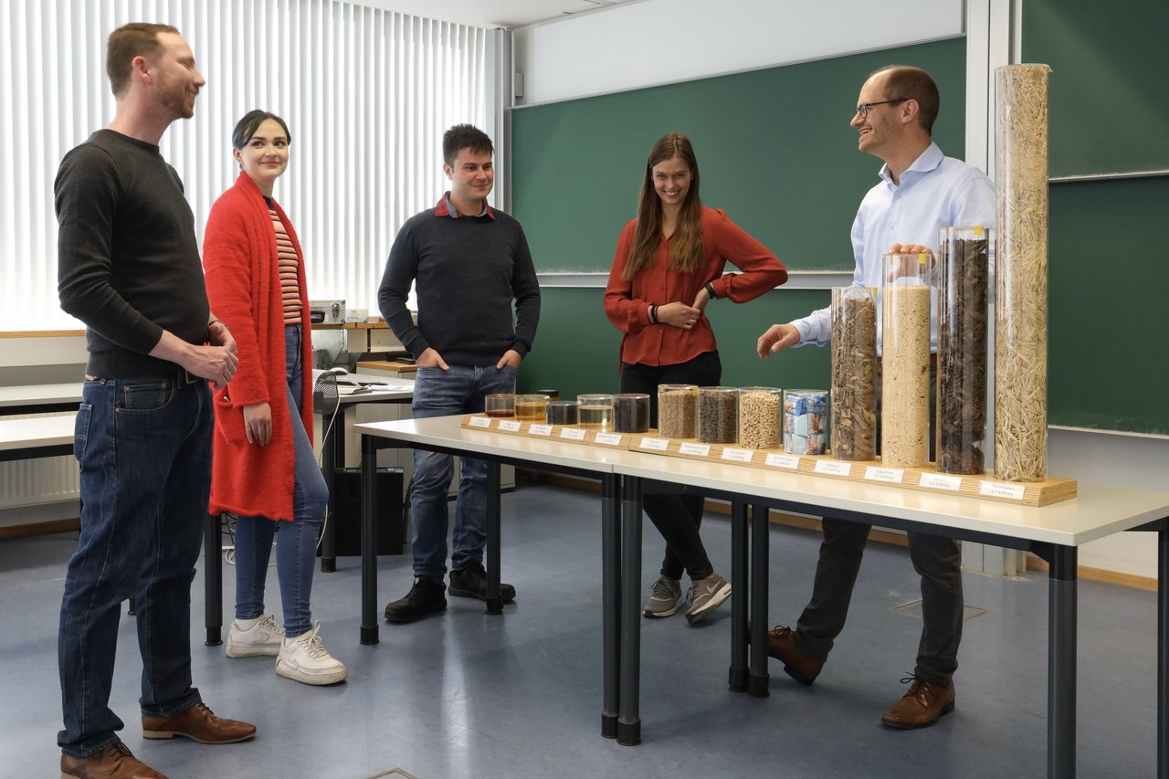 Professor zeigt einer Gruppe von Studierenden verschiedene Materialien, um die Wattstunden pro Kilogramm, die aus den jeweiligen Materialien gewonnen werden können, darzustellen
